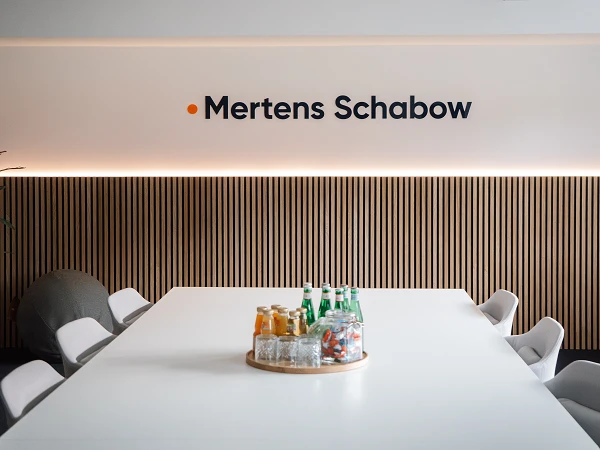 Ein modern gestalteter Besprechungsraum mit einem langen weißen Tisch, umgeben von weißen Stühlen. Auf dem Tisch befindet sich ein runder Holzuntersetzer mit verschiedenen Getränken und Gläsern. An der Wand steht der Schriftzug "Mertens Schabow" in großen Buchstaben, unterstrichen von einer warmen Beleuchtung.