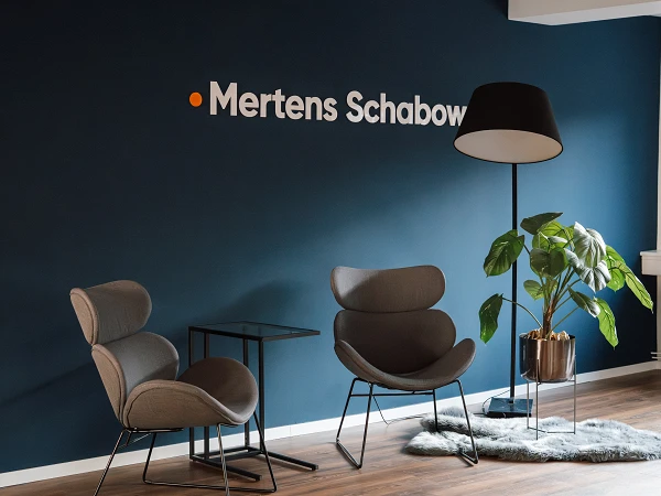 Ein modern gestalteter Empfangsbereich mit einem dunkelblauen Wandhintergrund, auf dem der Schriftzug "Mertens Schabow" in weiß und orange zu sehen ist. Vor der Wand stehen zwei elegante, graue Sessel, ein kleiner Tisch und eine moderne Stehlampe. Eine grüne Pflanze steht in einem goldenen Topf daneben.