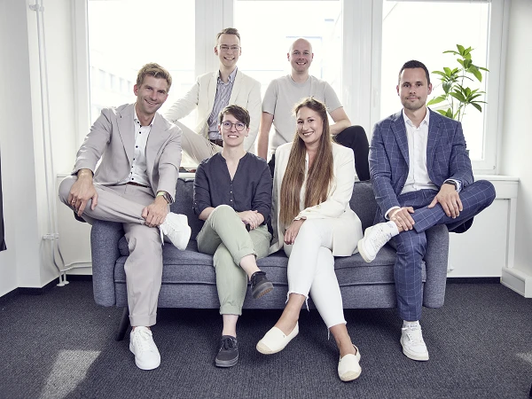 Sechs Personen sitzen und stehen in einem modernen Büroraum auf einem Sofa, lächelnd in die Kamera; sie tragen verschiedene formelle Kleidung in hellen Farben und sind gut gekleidet. Im Hintergrund sind große Fenster und eine Pflanze zu sehen.