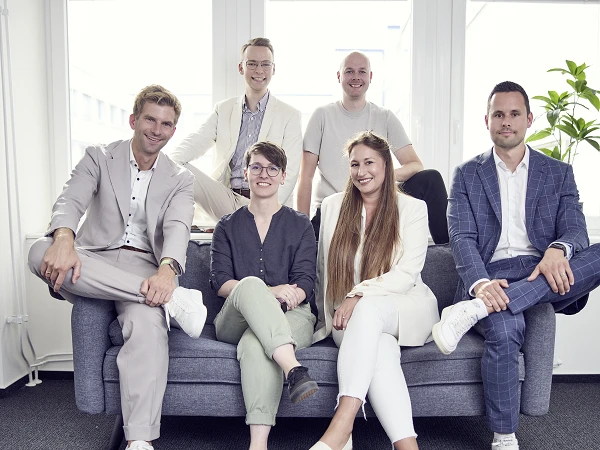 Sechs Personen sitzen und stehen in einem modernen Büroraum auf einem Sofa, lächelnd in die Kamera; sie tragen verschiedene formelle Kleidung in hellen Farben und sind gut gekleidet. Im Hintergrund sind große Fenster und eine Pflanze zu sehen.