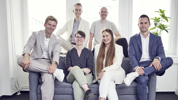 Sechs Personen sitzen und stehen in einem modernen Büroraum auf einem Sofa, lächelnd in die Kamera; sie tragen verschiedene formelle Kleidung in hellen Farben und sind gut gekleidet. Im Hintergrund sind große Fenster und eine Pflanze zu sehen.