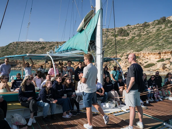Eine Gruppe von Menschen sitzt auf einem Segelboot, während zwei Männer im Vordergrund sprechen. Der Hintergrund zeigt eine Küstenlandschaft mit grünen Hügeln und klarem blauen Wasser. Einige Personen halten Tassen oder Getränke in den Händen.