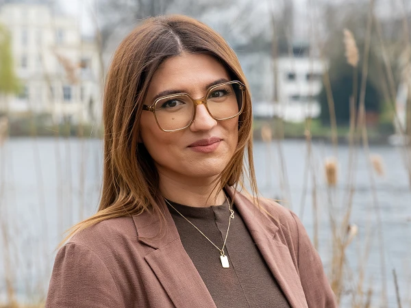 Eine junge Frau mit langen, braunen Haaren und einer bräunlichen Jacke steht am Ufer eines Gewässers, umgeben von einer natürlichen Landschaft mit Schilfrohr im Vordergrund und einer sanften Bebauung im Hintergrund.