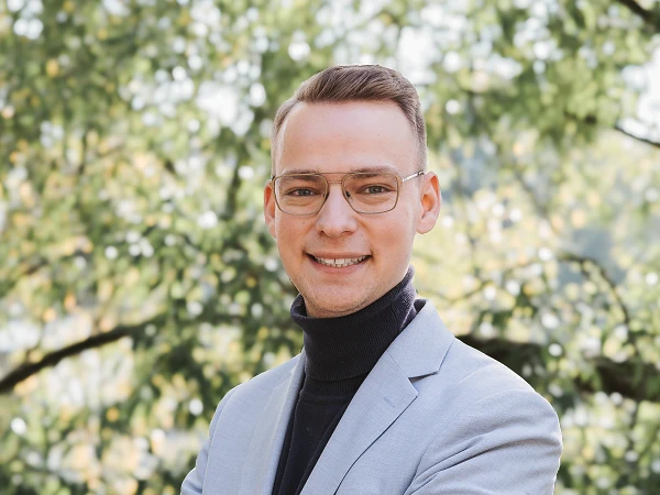Ein junger Mann in einem grauen Blazer und einem schwarzen Rollkragenpullover steht mit verschränkten Armen vor einem Hintergrund aus grünen Bäumen und einem weißen Himmel, während er lächelt.