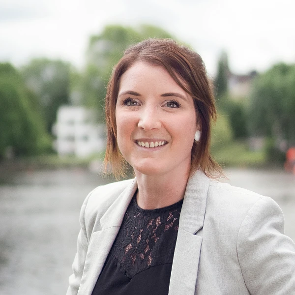Frau mit Blazer und Spitzentop, stehend an einem Flussufer, im Hintergrund grüne Bäume