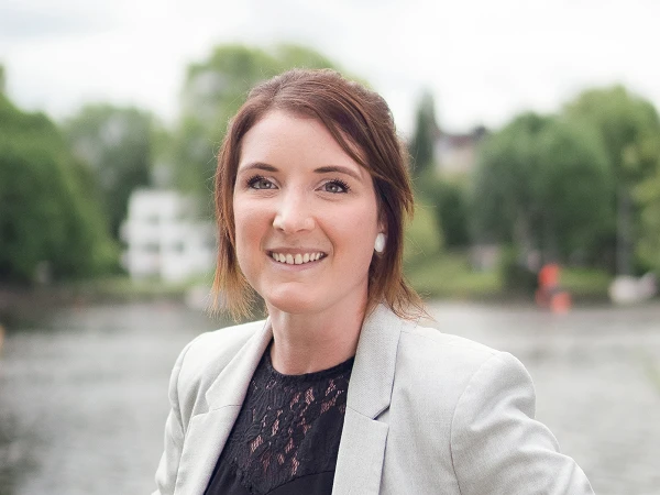Frau mit Blazer und Spitzentop, stehend an einem Flussufer, im Hintergrund grüne Bäume