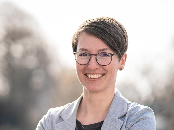 Frau mit kurzem dunklem Haar und Brille, trägt grauen Blazer, lächelt in die Kamera vor unscharfem Hintergrund