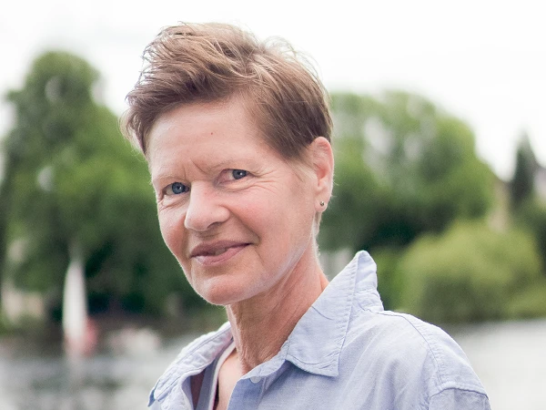 Frau mit kurzem Haar im blauen Hemd, vor Flusslandschaft mit grünen Bäumen