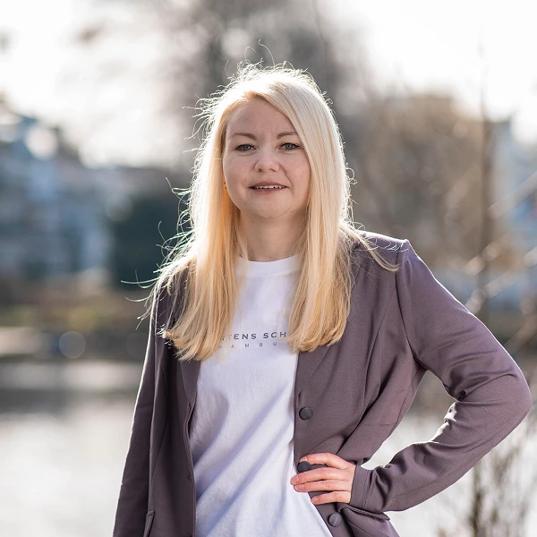 Frau mit langen blonden Haaren im hellvioletten Blazer und weißem T-Shirt, stehend am Wasser