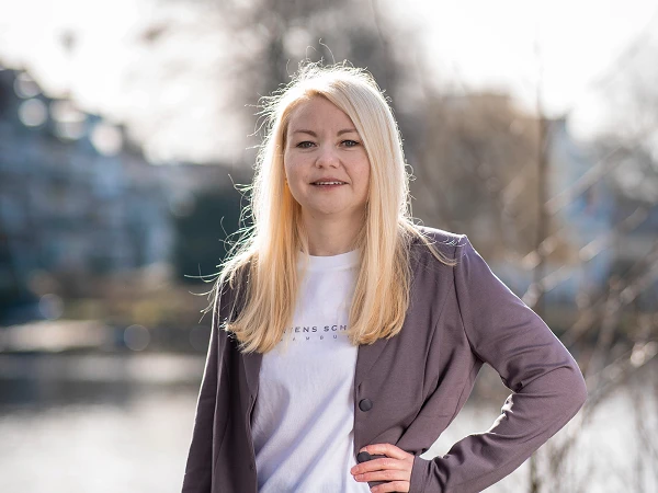 Frau mit langen blonden Haaren im hellvioletten Blazer und weißem T-Shirt, stehend am Wasser