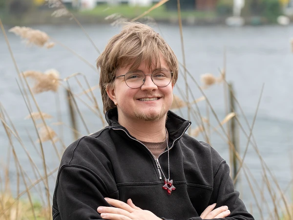 Junger Mann mit Brille und einem lächelnden Gesicht steht mit verschränkten Armen am Ufer eines Gewässers, umgeben von hohen Gräsern und Schilfpflanzen.