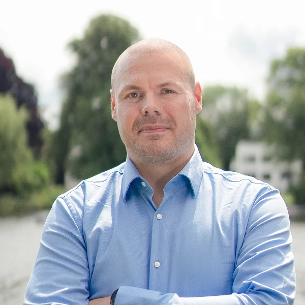 Mann im blauen Hemd mit verschränkten Armen vor See und Bäumen