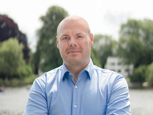 Mann im blauen Hemd mit verschränkten Armen vor See und Bäumen