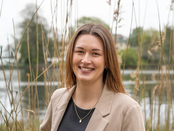 Junge Frau mit dunklem Oberteil und beigem Blazer vor Schilf und See
