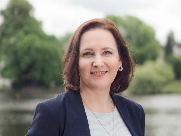 Frau mit braunen Haaren im Blazer vor See und Bäumen