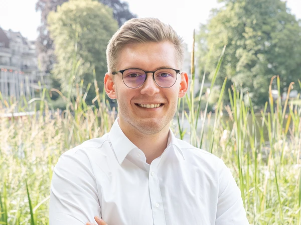 Ein junger Mann mit Brille und einem weißen Hemd steht lächelnd mit verschränkten Armen vor einer grünen Wiese, umgeben von Bäumen und einem Fluss im Hintergrund.