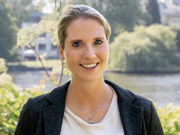 Eine lächelnde Frau mit blonden Haaren steht vor einem ruhigen Gewässer, umgeben von grünen Pflanzen und Bäumen, die im Hintergrund sichtbar sind. Sie trägt einen dunkelgrauen Blazer über einem hellen Oberteil.