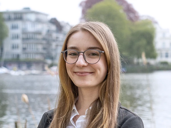 Junge Frau mit Brille im schwarzen Blazer vor See und Gebäuden