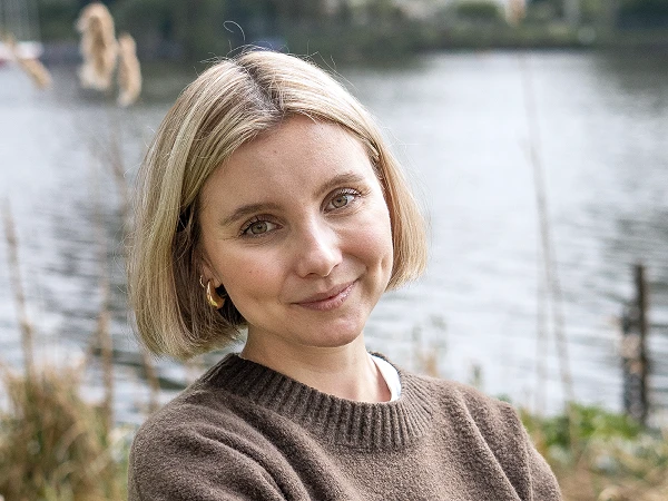 Frau mit blondem Bob und braunem Pullover vor Wasser