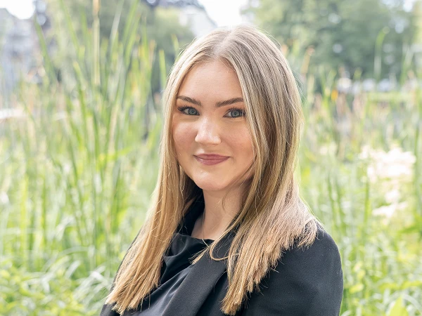 Eine junge Frau mit langen, braunen Haaren steht vor einer grünen, blühenden Umgebung mit hohen Gräsern und Pflanzen. Sie trägt einen schwarzen Blazer und lächelt in die Kamera.