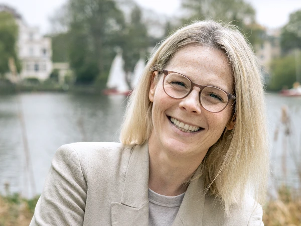 Lächelnde Frau mit Brille und blondem Haar im beigen Blazer vor See