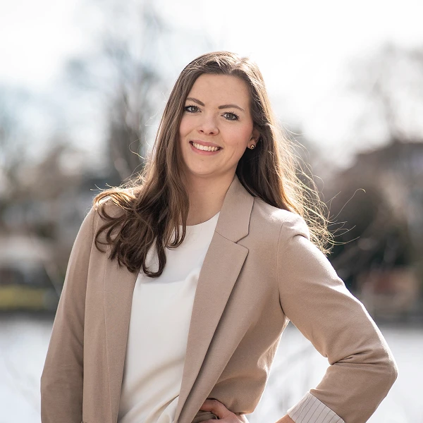 Frau mit langen braunen Haaren im beigen Blazer vor Wasserlandschaft