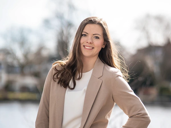 Frau mit langen braunen Haaren im beigen Blazer vor Wasserlandschaft