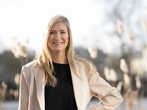 Frau mit langen blonden Haaren im hellen Blazer vor Schilf und Wasser