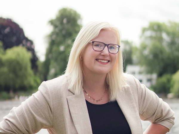 Frau mit blonden Haaren, Brille und beigefarbenem Blazer am Wasser