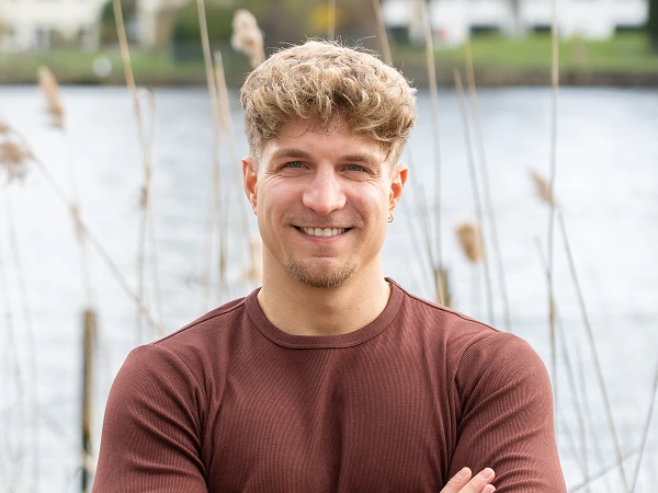 Ein junger Mann mit geföhnten, blonden Locken steht mit verschränkten Armen vor einem Gewässer, umgeben von hohen Gräsern und einer natürlichen Umgebung. Er lächelt freundlich in die Kamera.