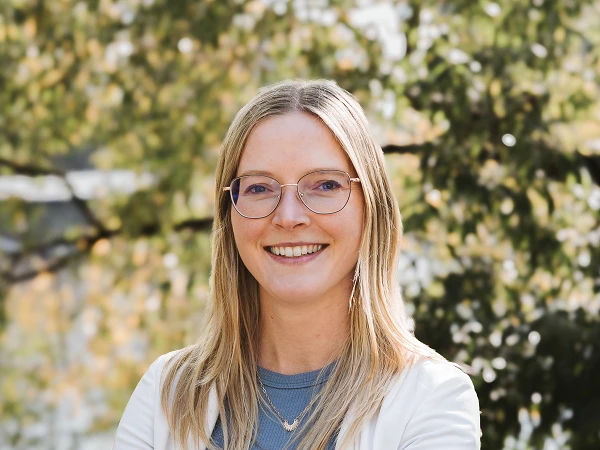 Eine lächelnde Frau mit langen, blonden Haaren und Brille steht vor einer grünen, baumbestandenen Kulisse. Sie trägt ein graues Oberteil und eine weiße Jacke und wirkt entspannt und freundlichen. Im Hintergrund ist unscharf ein Wasserbereich zu erkennen.