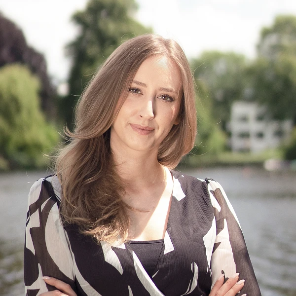 Frau mit langen hellbraunen Haaren in gemusterter Bluse vor Wasser