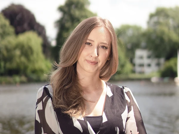 Frau mit langen hellbraunen Haaren in gemusterter Bluse vor Wasser