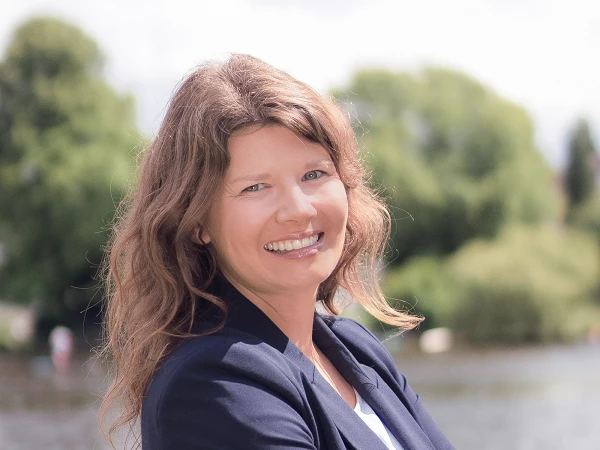 Frau mit dunkelblondem Haar im Blazer, lächelt vor Flusslandschaft