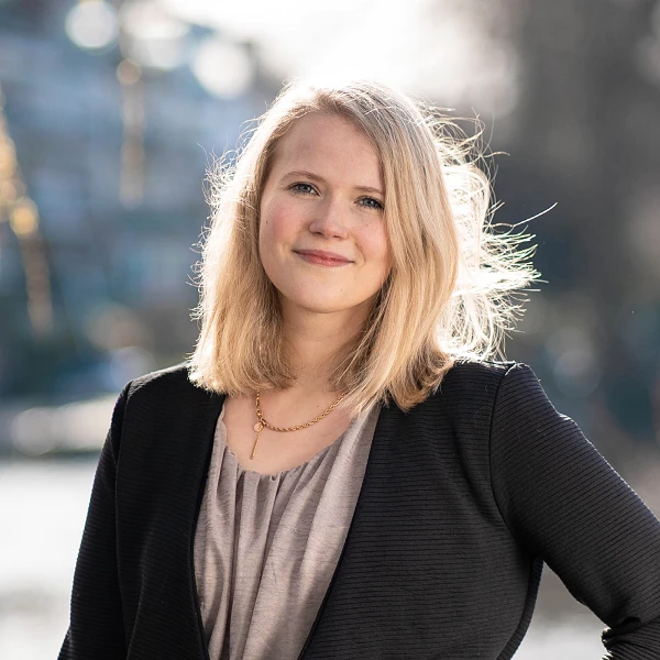 Frau mit dunkelblondem Haar im Blazer, lächelt vor Flusslandschaft
