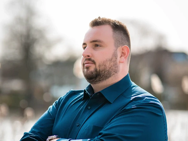 Mann mit Brille und blauem Hemd verschränkt die Arme am Wasser