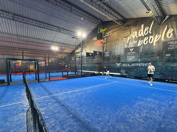 Ein Innenraum eines Padel-Tennisplatzes mit blauen Böden, in dem zwei Spieler aktiv ein Spiel spielen. Im Hintergrund sind die Wände mit Werbeplakaten und Logos versehen.