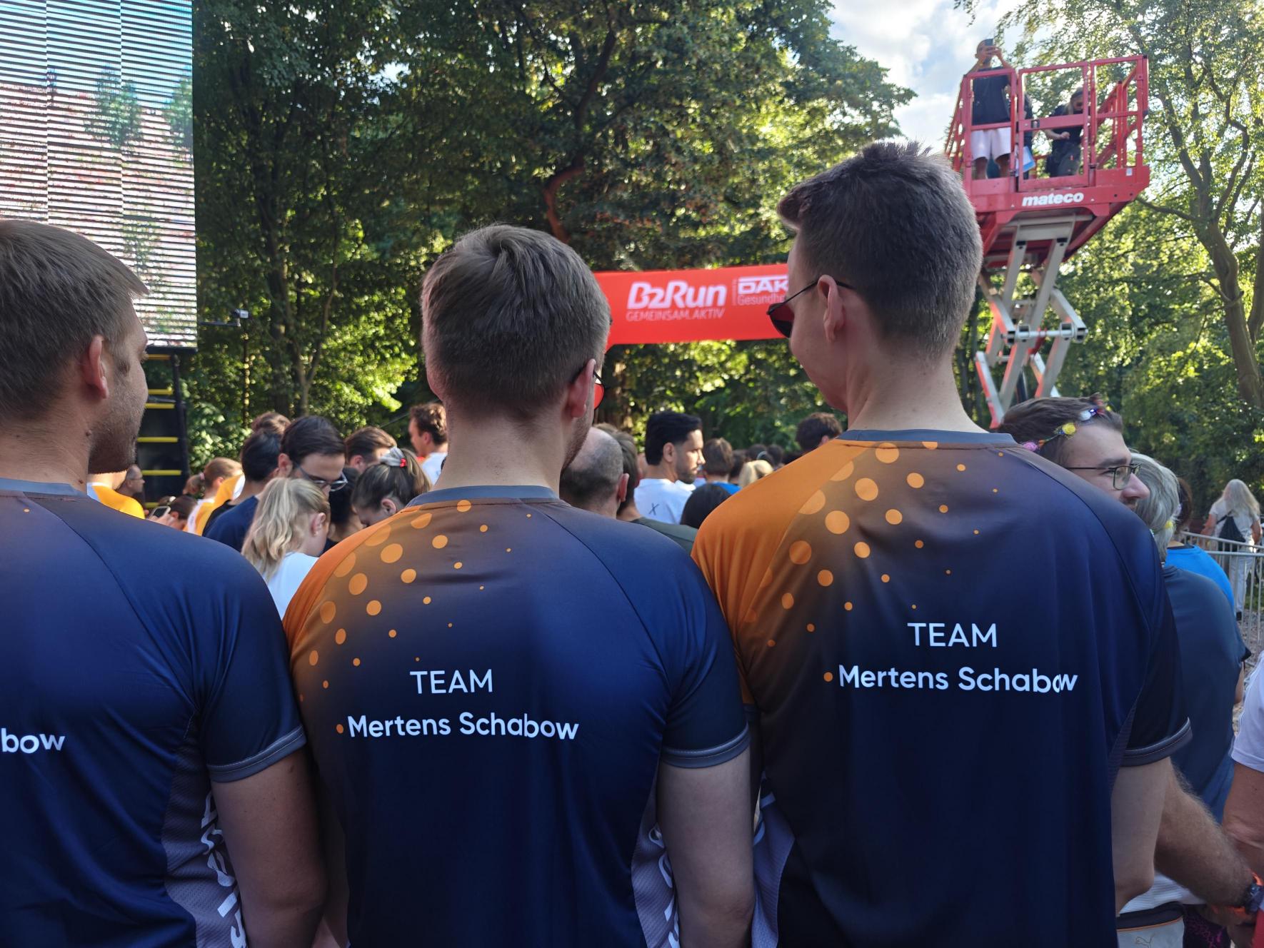 Drei Personen stehen mit dem Rücken zur Kamera in sportlichen Trikots, auf denen der Text „TEAM Mertens Schabow“ steht, während sie sich auf ein Ereignis im Freien vorbereiten. Im Hintergrund sind viele Menschen und eine große Bühne zu sehen.