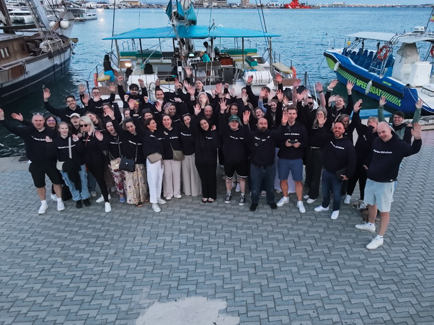 Eine große Gruppe von Menschen steht auf einem Steg am Wasser, sie tragen schwarze Sweatshirts und winken in die Kamera. Im Hintergrund sind Boote und ein Hafen zu sehen.