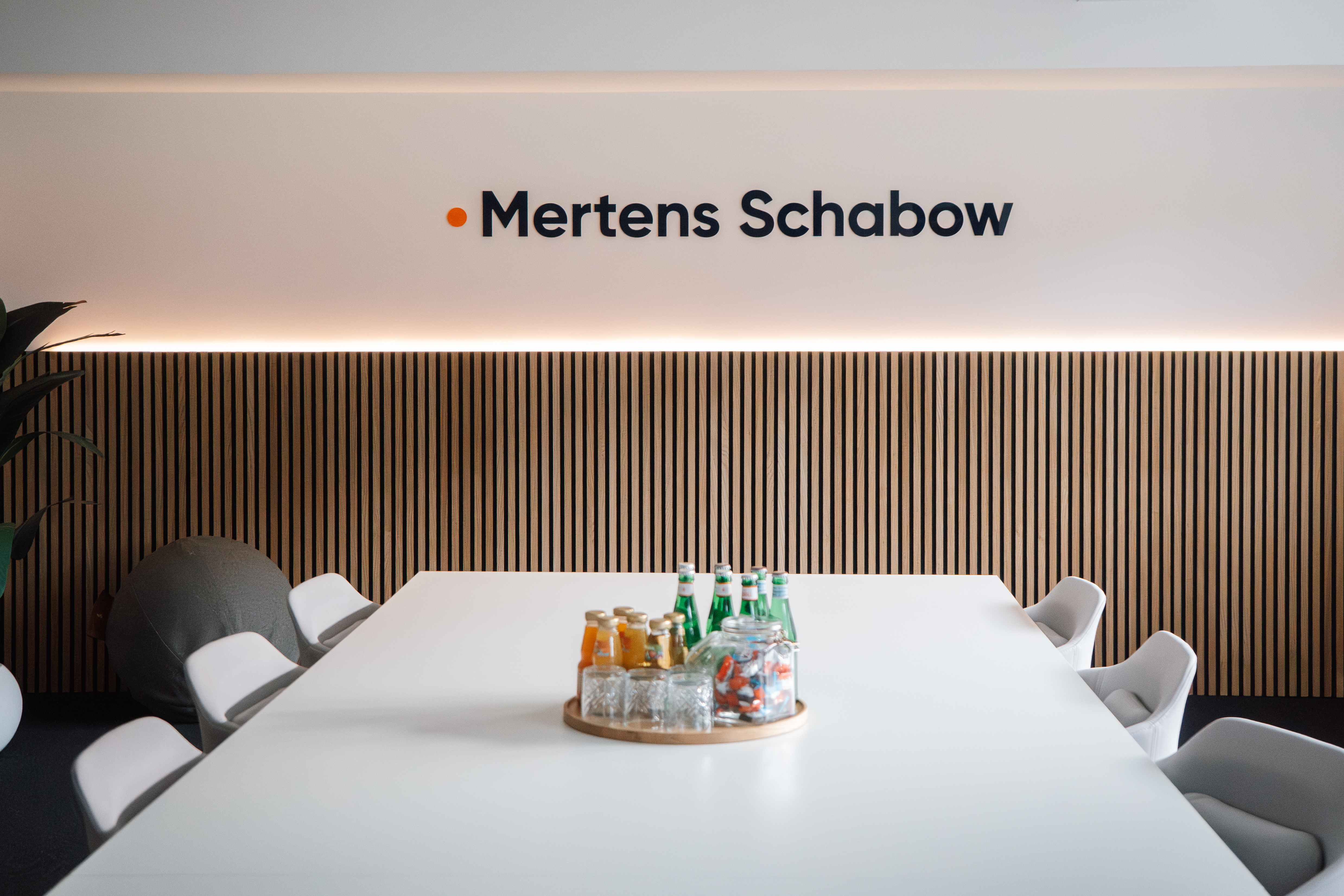 Ein modern gestalteter Besprechungsraum mit einem langen weißen Tisch, umgeben von weißen Stühlen. Auf dem Tisch befindet sich ein runder Holzuntersetzer mit verschiedenen Getränken und Gläsern. An der Wand steht der Schriftzug "Mertens Schabow" in großen Buchstaben, unterstrichen von einer warmen Beleuchtung.
