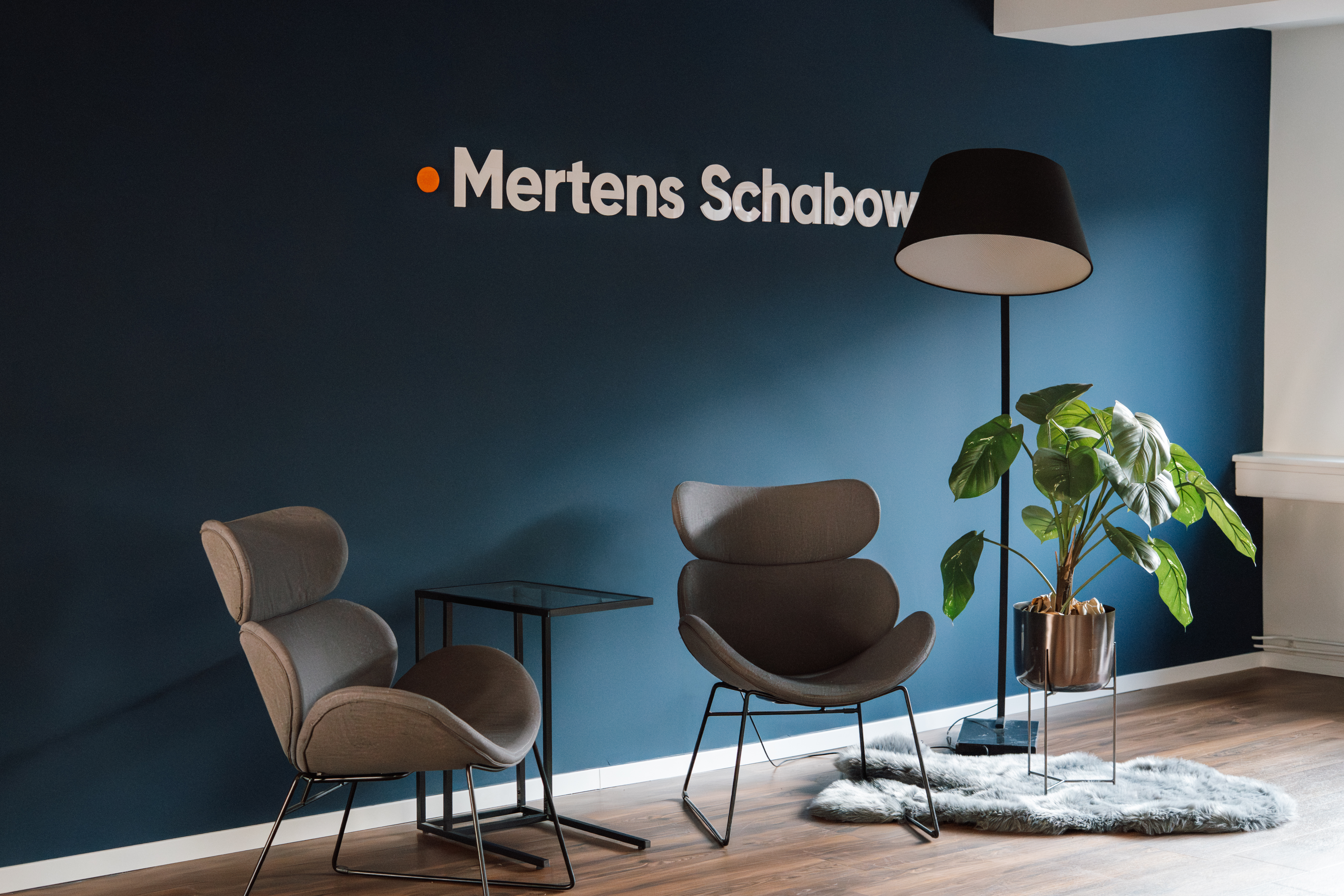 Ein modern gestalteter Empfangsbereich mit einem dunkelblauen Wandhintergrund, auf dem der Schriftzug "Mertens Schabow" in weiß und orange zu sehen ist. Vor der Wand stehen zwei elegante, graue Sessel, ein kleiner Tisch und eine moderne Stehlampe. Eine grüne Pflanze steht in einem goldenen Topf daneben.