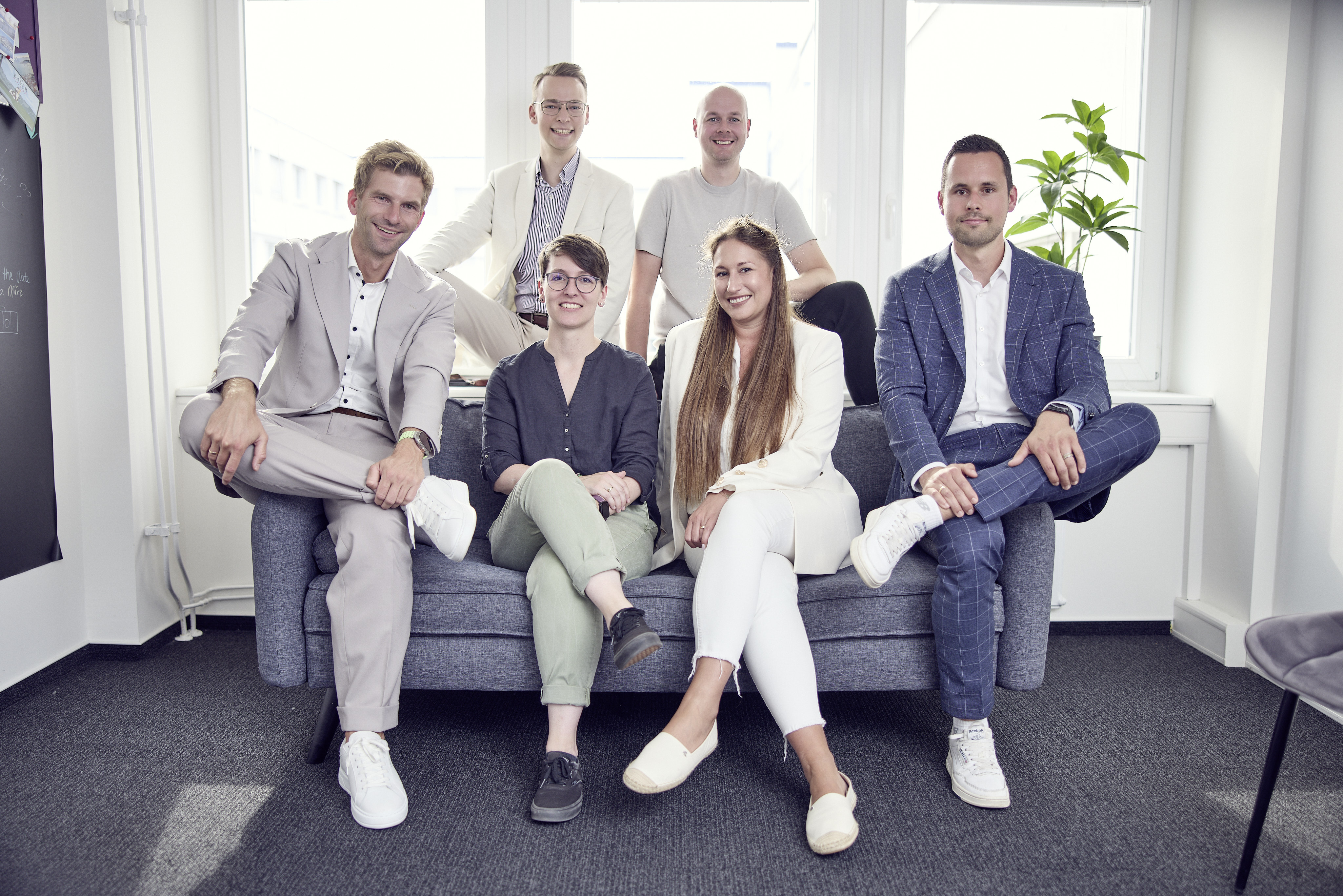 Sechs Personen sitzen und stehen in einem modernen Büroraum auf einem Sofa, lächelnd in die Kamera; sie tragen verschiedene formelle Kleidung in hellen Farben und sind gut gekleidet. Im Hintergrund sind große Fenster und eine Pflanze zu sehen.