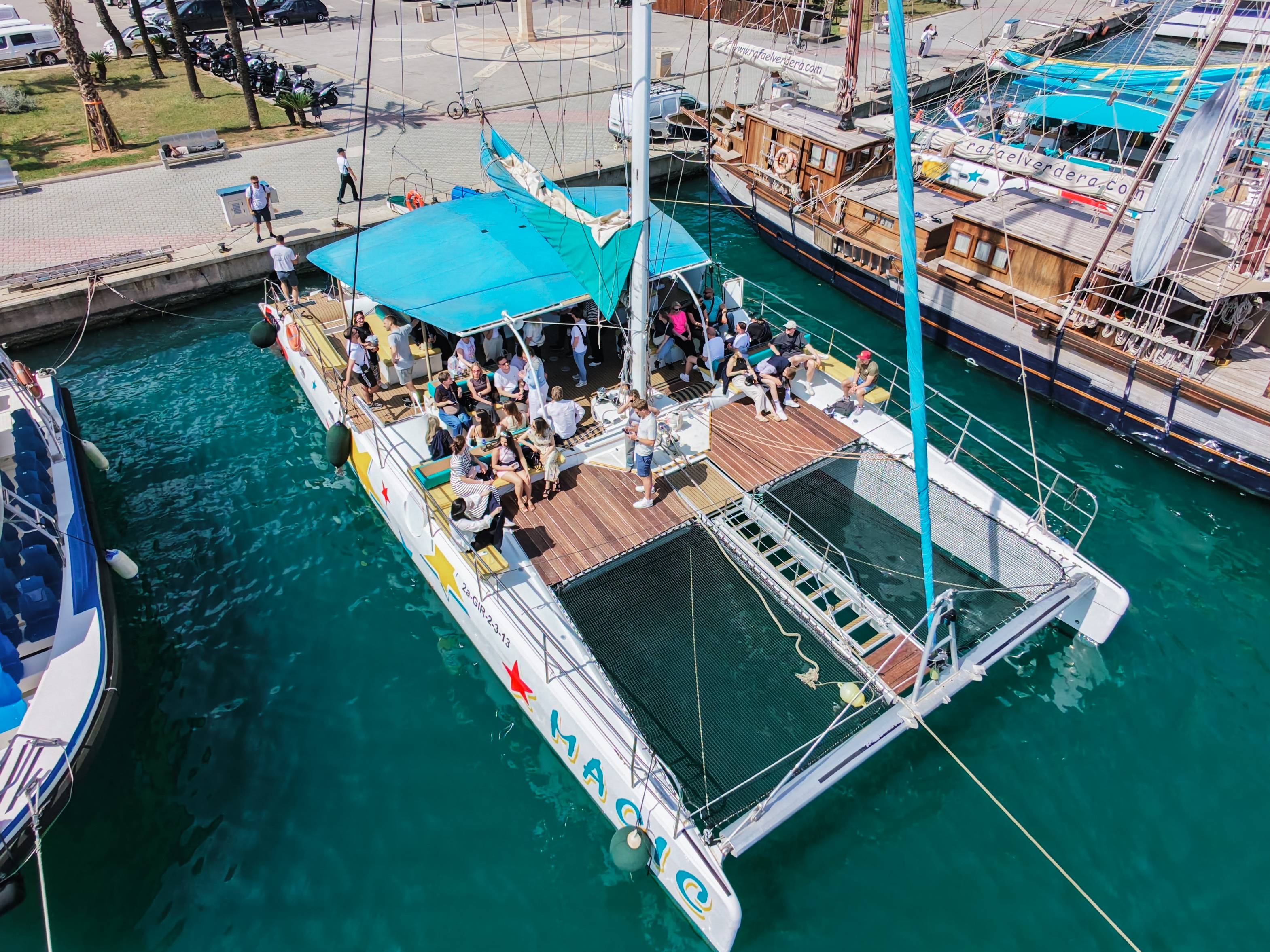 Ein Katamaran mit blauer Plane liegt im Hafen, umgeben von anderen Booten. Auf dem Deck sitzen zahlreiche Personen, die sich entspannen und die Aussicht genießen. Im Hintergrund sind einige Palmen und Hafenanlagen zu sehen.