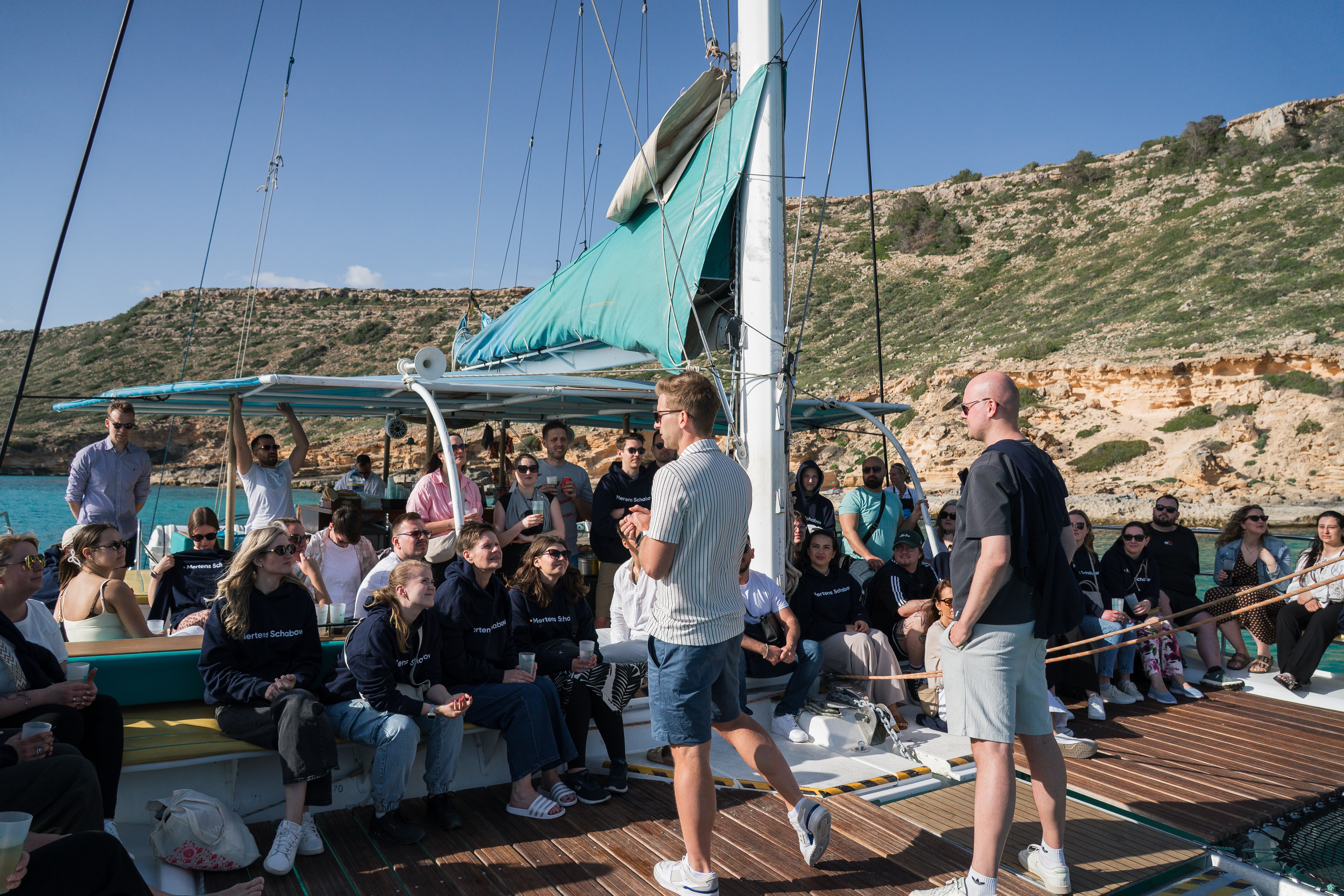 Eine Gruppe von Menschen sitzt auf einem Segelboot, während zwei Männer im Vordergrund sprechen. Der Hintergrund zeigt eine Küstenlandschaft mit grünen Hügeln und klarem blauen Wasser. Einige Personen halten Tassen oder Getränke in den Händen.