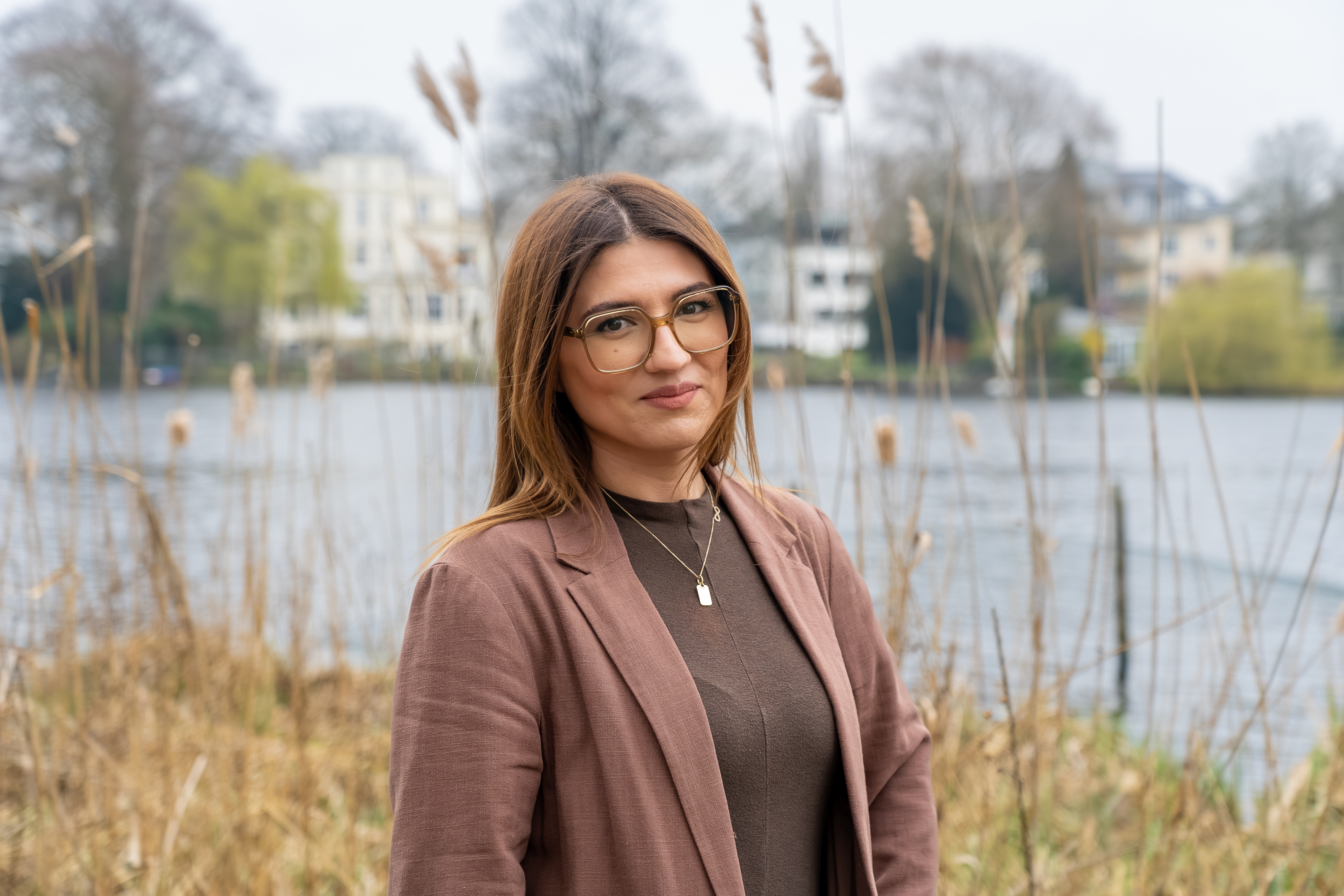 Eine junge Frau mit langen, braunen Haaren und einer bräunlichen Jacke steht am Ufer eines Gewässers, umgeben von einer natürlichen Landschaft mit Schilfrohr im Vordergrund und einer sanften Bebauung im Hintergrund.