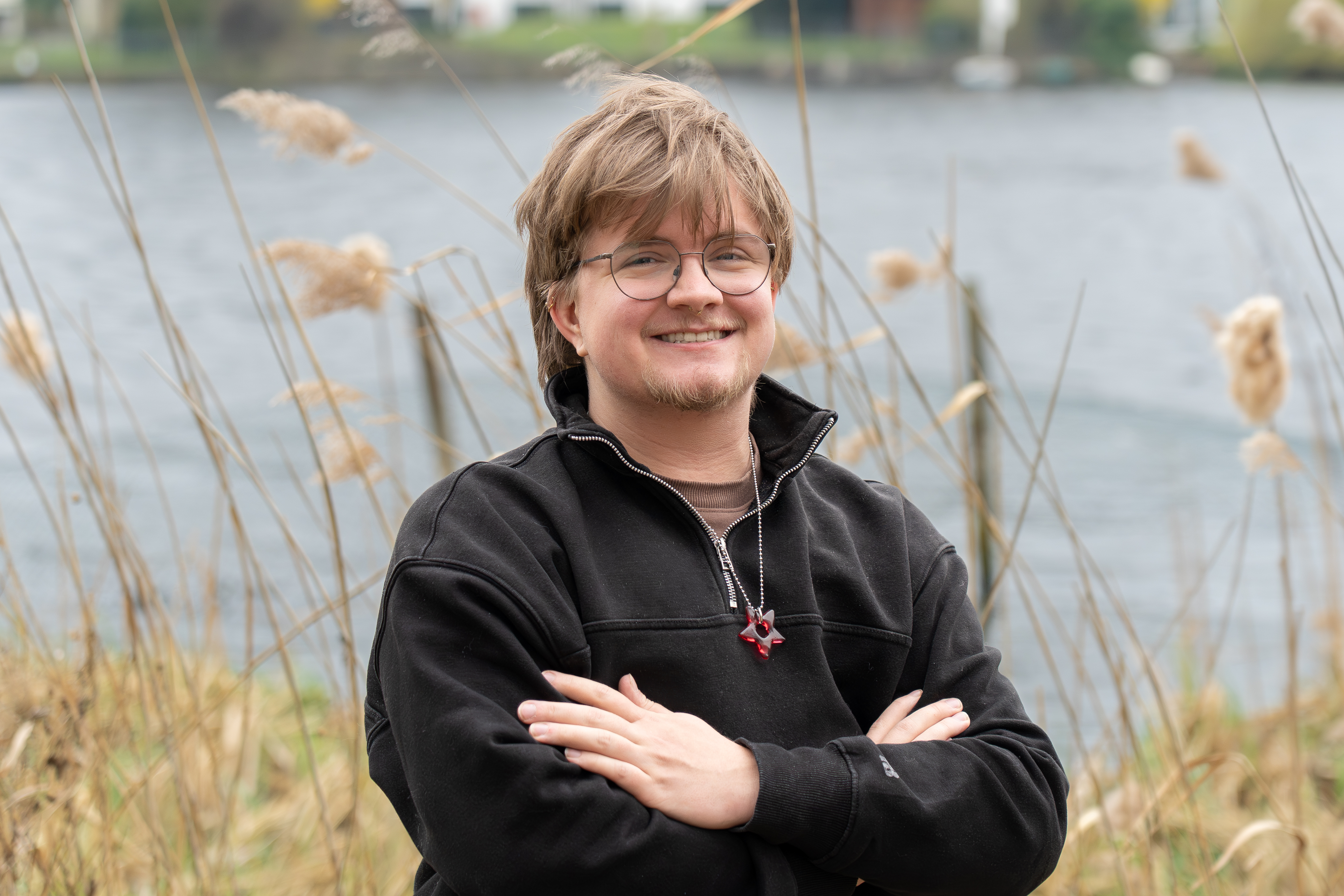 Junger Mann mit Brille und einem lächelnden Gesicht steht mit verschränkten Armen am Ufer eines Gewässers, umgeben von hohen Gräsern und Schilfpflanzen.