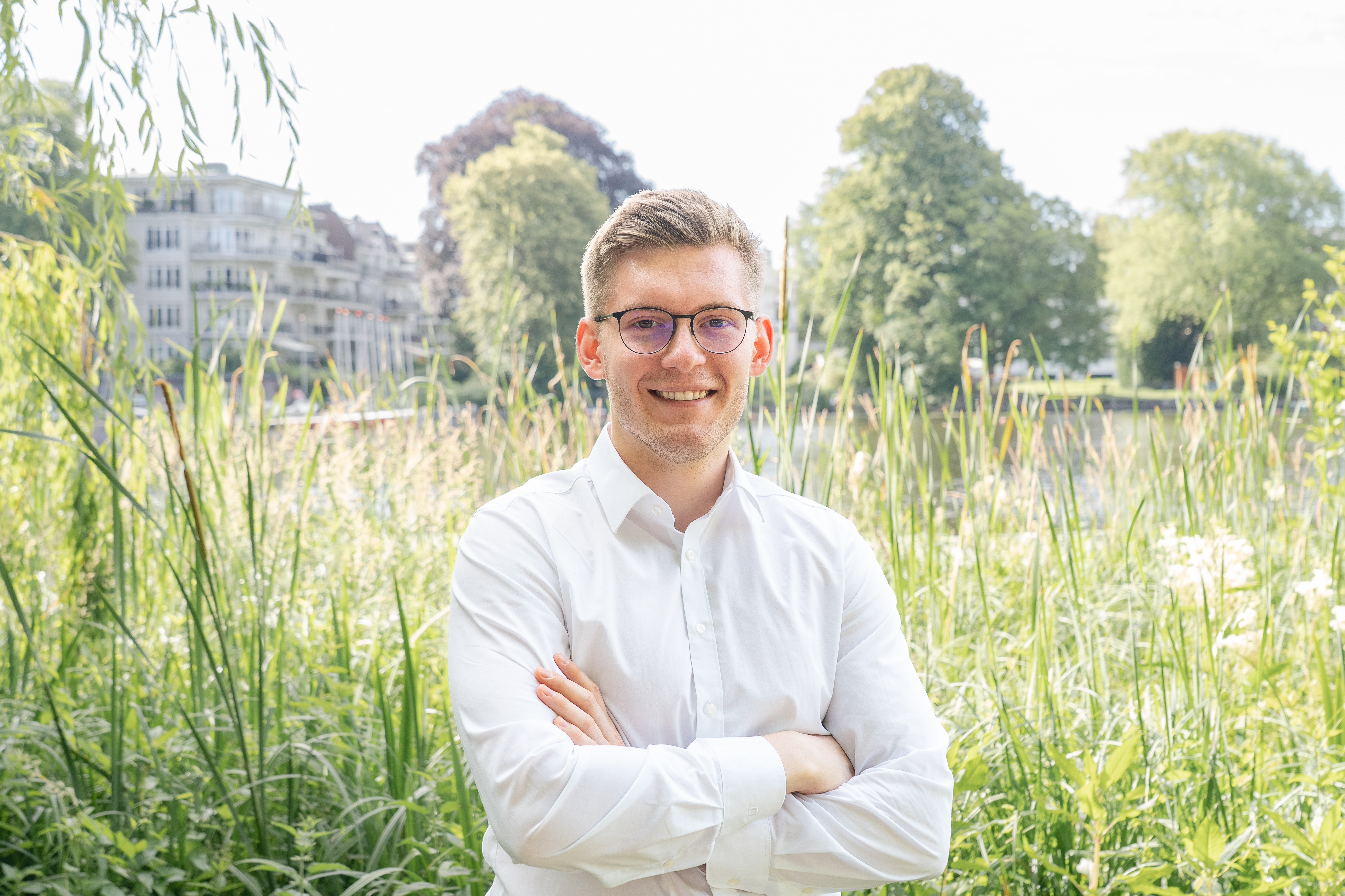 Ein junger Mann mit Brille und einem weißen Hemd steht lächelnd mit verschränkten Armen vor einer grünen Wiese, umgeben von Bäumen und einem Fluss im Hintergrund.