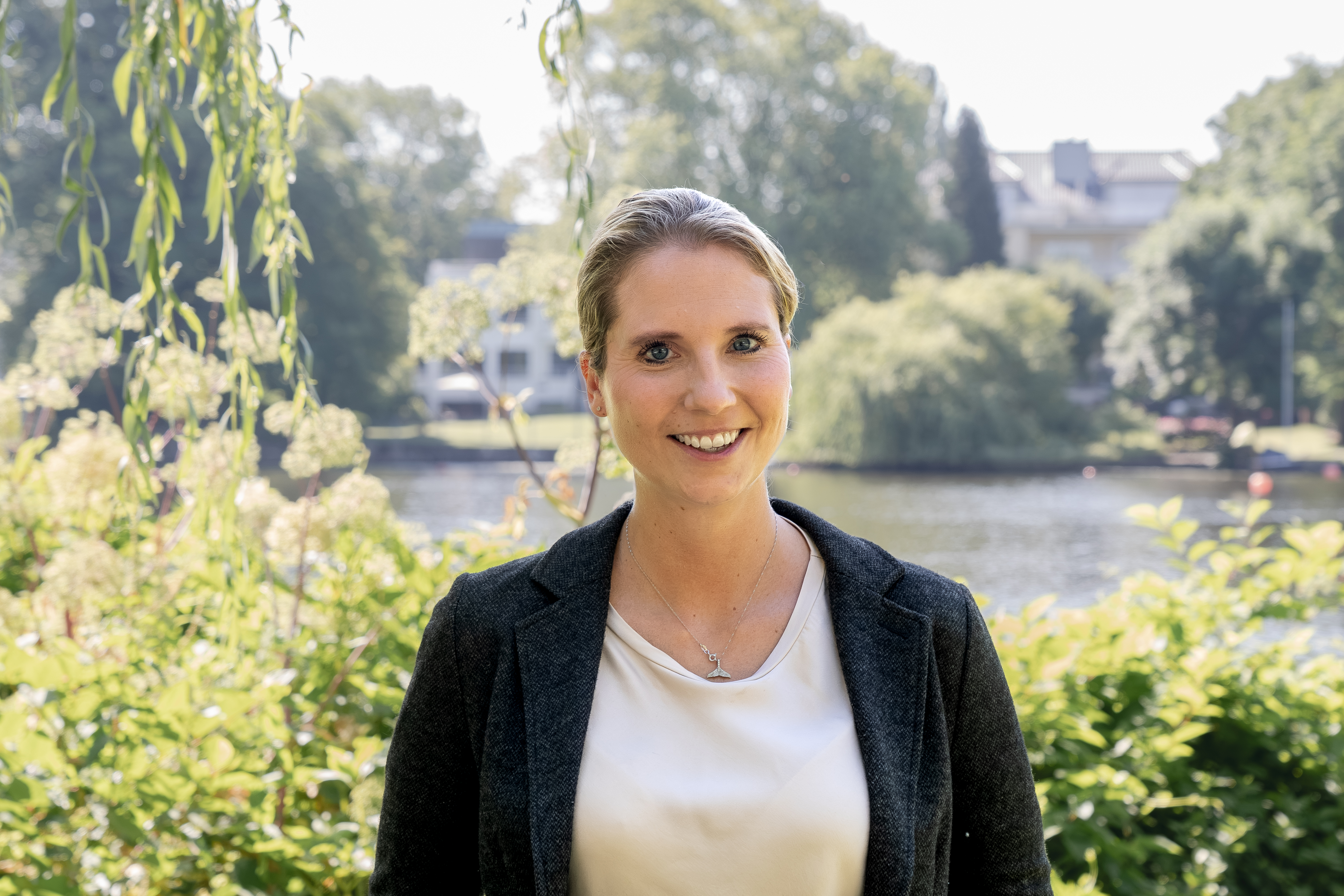 Eine lächelnde Frau mit blonden Haaren steht vor einem ruhigen Gewässer, umgeben von grünen Pflanzen und Bäumen, die im Hintergrund sichtbar sind. Sie trägt einen dunkelgrauen Blazer über einem hellen Oberteil.