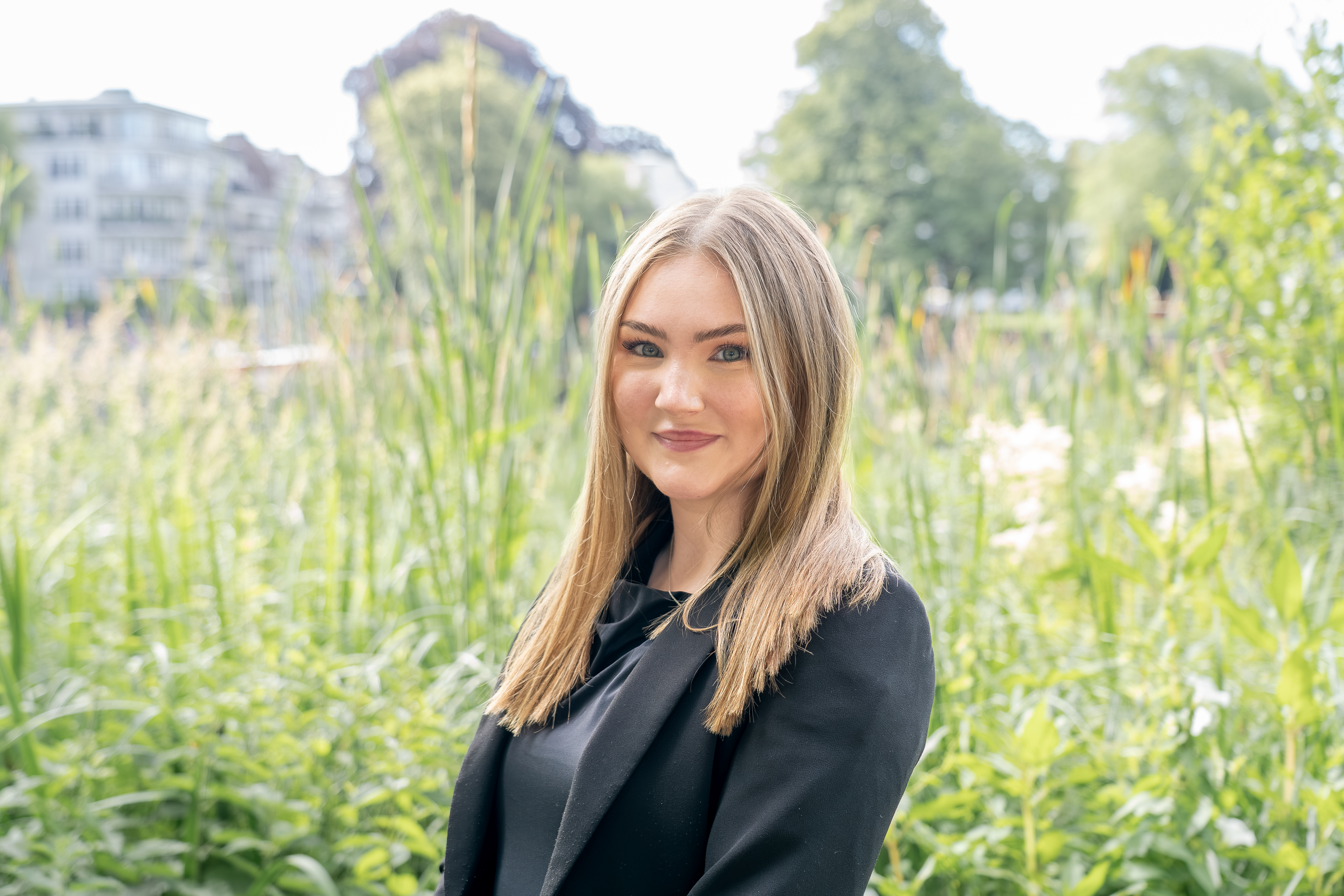 Eine junge Frau mit langen, braunen Haaren steht vor einer grünen, blühenden Umgebung mit hohen Gräsern und Pflanzen. Sie trägt einen schwarzen Blazer und lächelt in die Kamera.