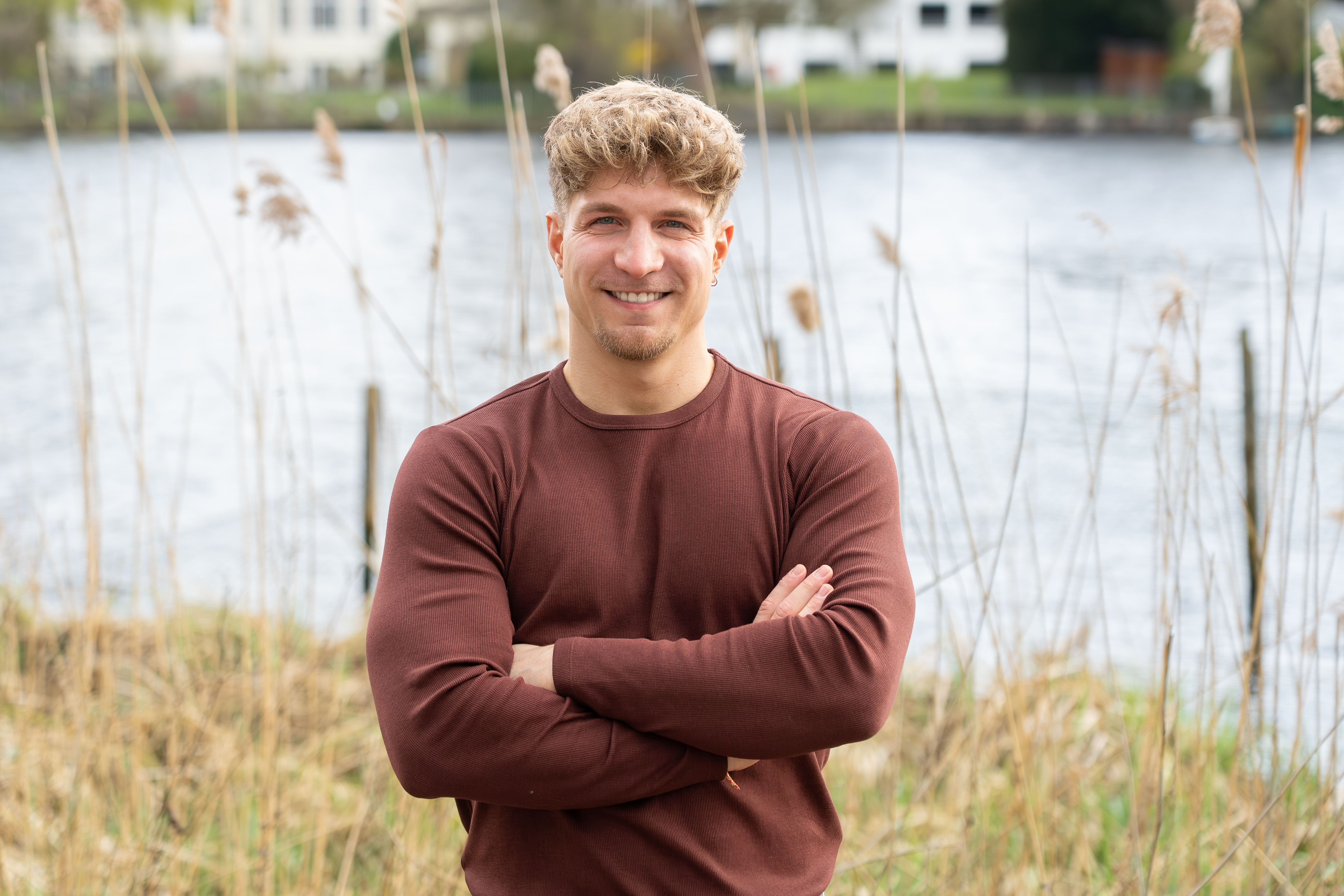 Ein junger Mann mit geföhnten, blonden Locken steht mit verschränkten Armen vor einem Gewässer, umgeben von hohen Gräsern und einer natürlichen Umgebung. Er lächelt freundlich in die Kamera.